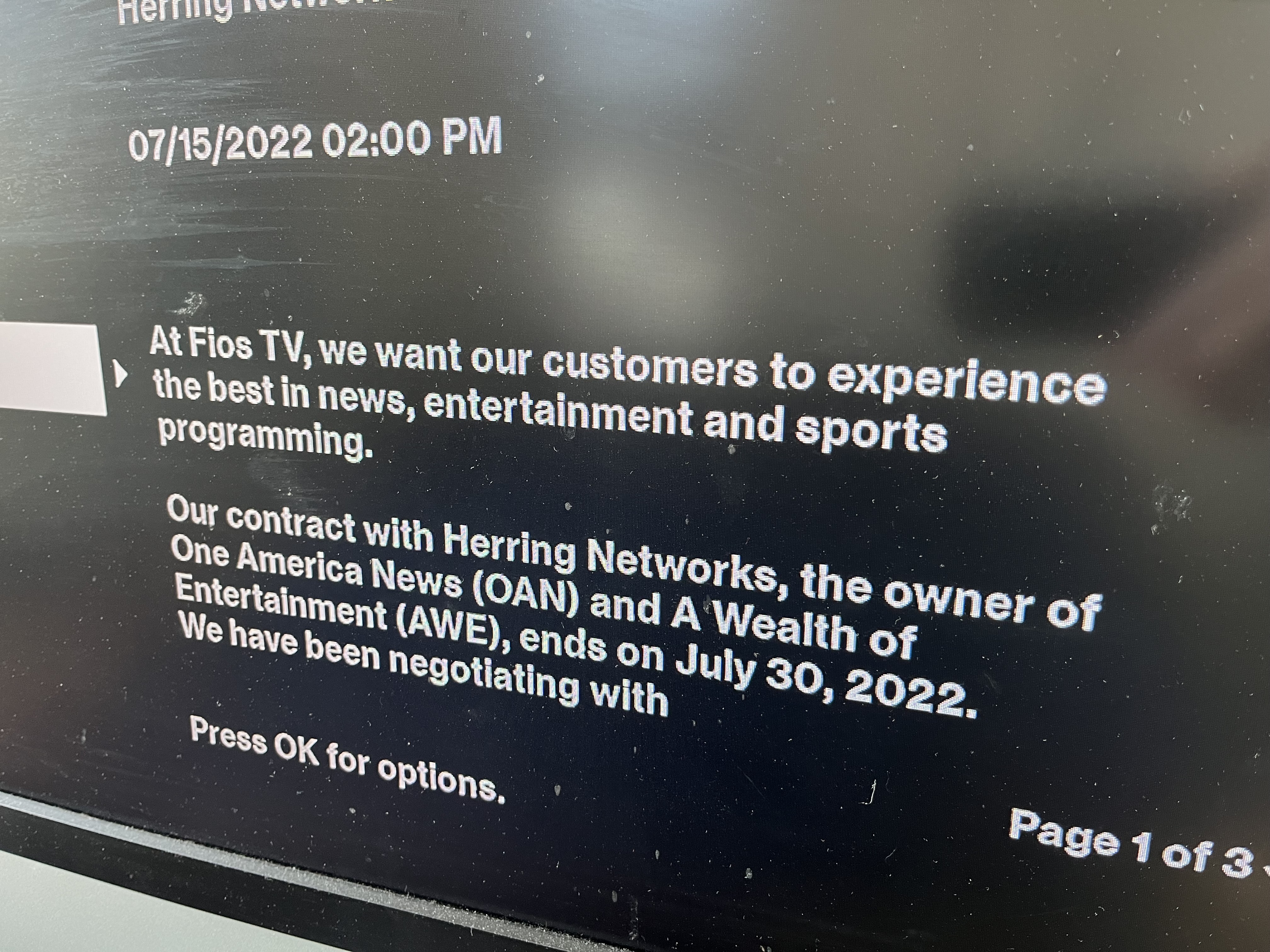 A photo of a TV displaying a message from Verizon Fios: "07/15/2022 02:00 PM At Fios TV, we want our customers to experience the best in news, entertainment, and sports programming. Our contract with Herring Networks, the owner of One America News (OAN) and A Wealth of Entertainment (AWE), ends on July 30, 2022. We have been negotiating with" Message continues on the next page.